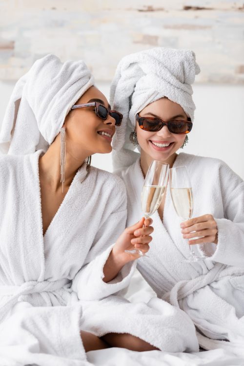 smiling interracial women in white soft bathrobes and trendy sunglasses clinking champagne glasses in bedroom. bath robes
