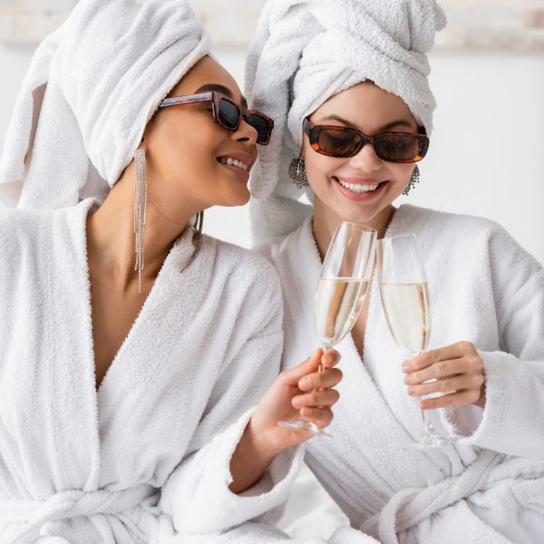 smiling interracial women in white soft bathrobes and trendy sunglasses clinking champagne glasses in bedroom. bath robes